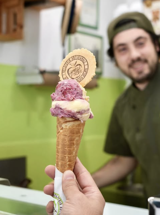 Two scoops of gelato are held with the Italian owner in the background 